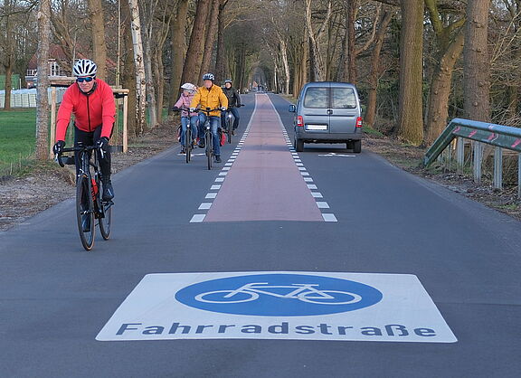 Eine Straße mit einem auf der Fahrbahn markierten Verkehrszeichen "Fahrradstraße". Dem Betrachter kommen mehrere Fahrradfahrer*innen entgegen. In der Mitte der Fahrbahn ist ein roter Streifen. Auf der rechten Fahrbahnseite fährt ein Auto vom Betrachter weg.