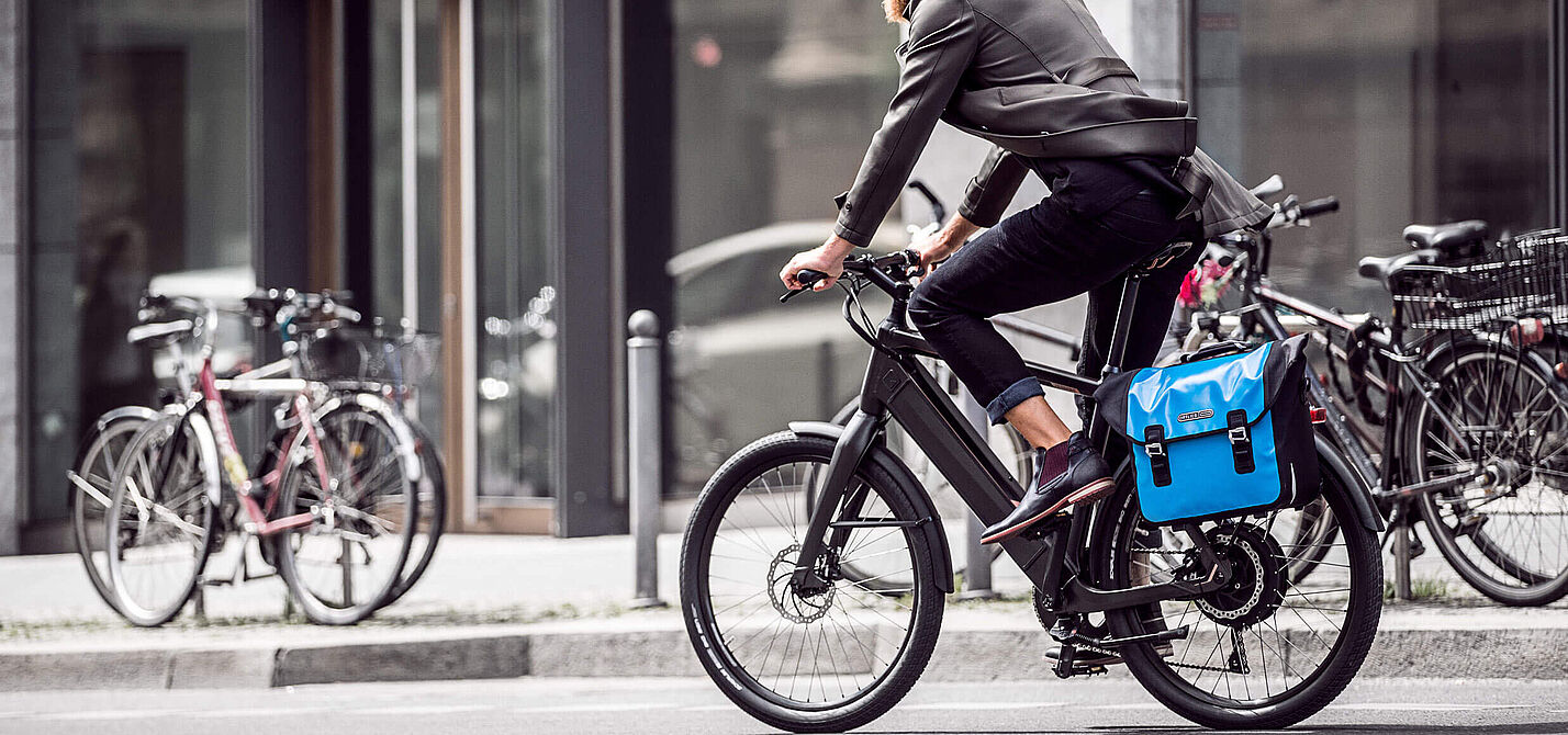 Berufspendler in der Stadt Ein Mann mit Bart und weißem Helm fährt auf einem modernen Elektrofahrrad durch eine städtische Straße. Er trägt Alltagskleidung (Jacke und Jeans) und hat eine leuchtend blaue Packtasche am Gepäckträger befestigt. Im Hintergrund sind Gebäude mit großen Glasfronten und parkende Fahrräder unscharf zu erkennen.